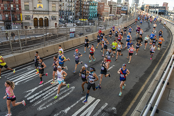 Peloton de coureurs sur une route urbaine de New York avec les bâtiments caractéristiques de la ville en arrière-plan.