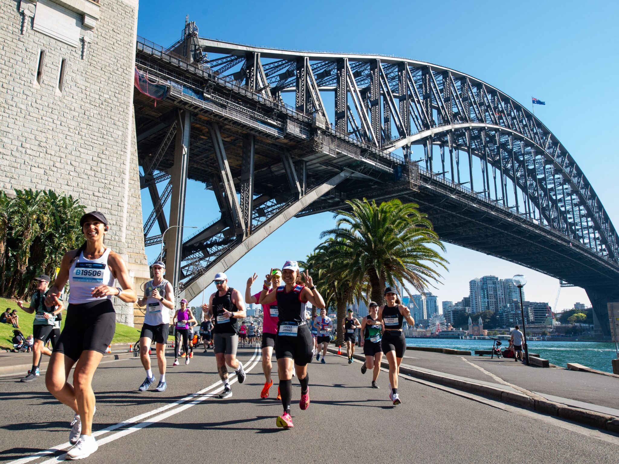 Coureurs du TCS Sydney Marathon passant sous le Sydney Harbour Bridge avec les palmiers et la baie en toile de fond.