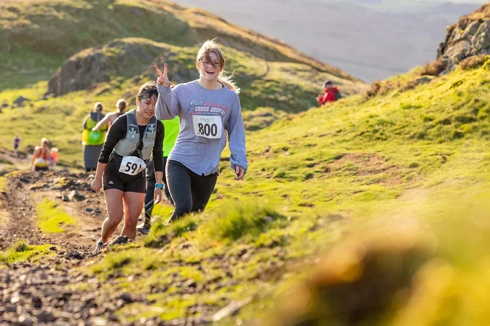 Coureuse souriante faisant le signe de la victoire sur un sentier ensoleillé des Ochil Hills.