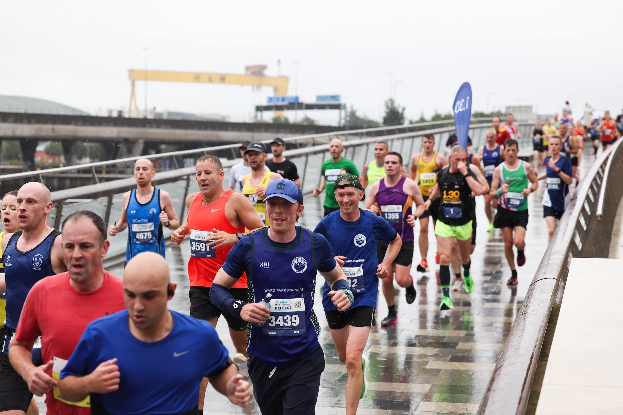 Coureurs du Belfast City Half Marathon traversant un pont sous la pluie avec les grues Harland & Wolff en arrière-plan.