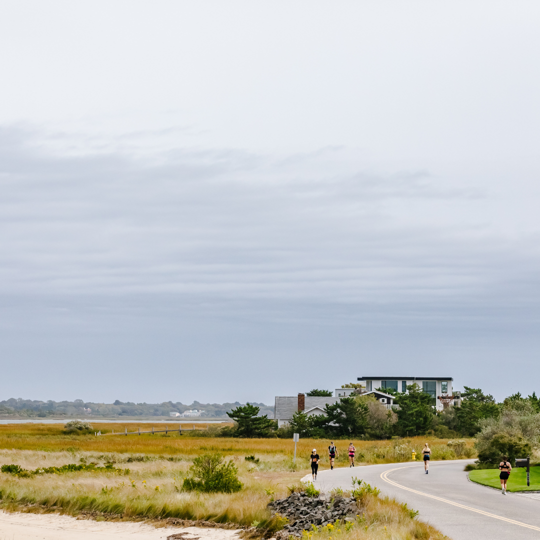 Coureurs sur le parcours du Hamptons Marathon longeant les marais côtiers et les maisons de plage typiques de Southampton.